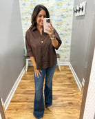 A woman takes a mirror selfie in a fitting room wearing a brown button-down shirt with rolled sleeves and blue flare jeans.