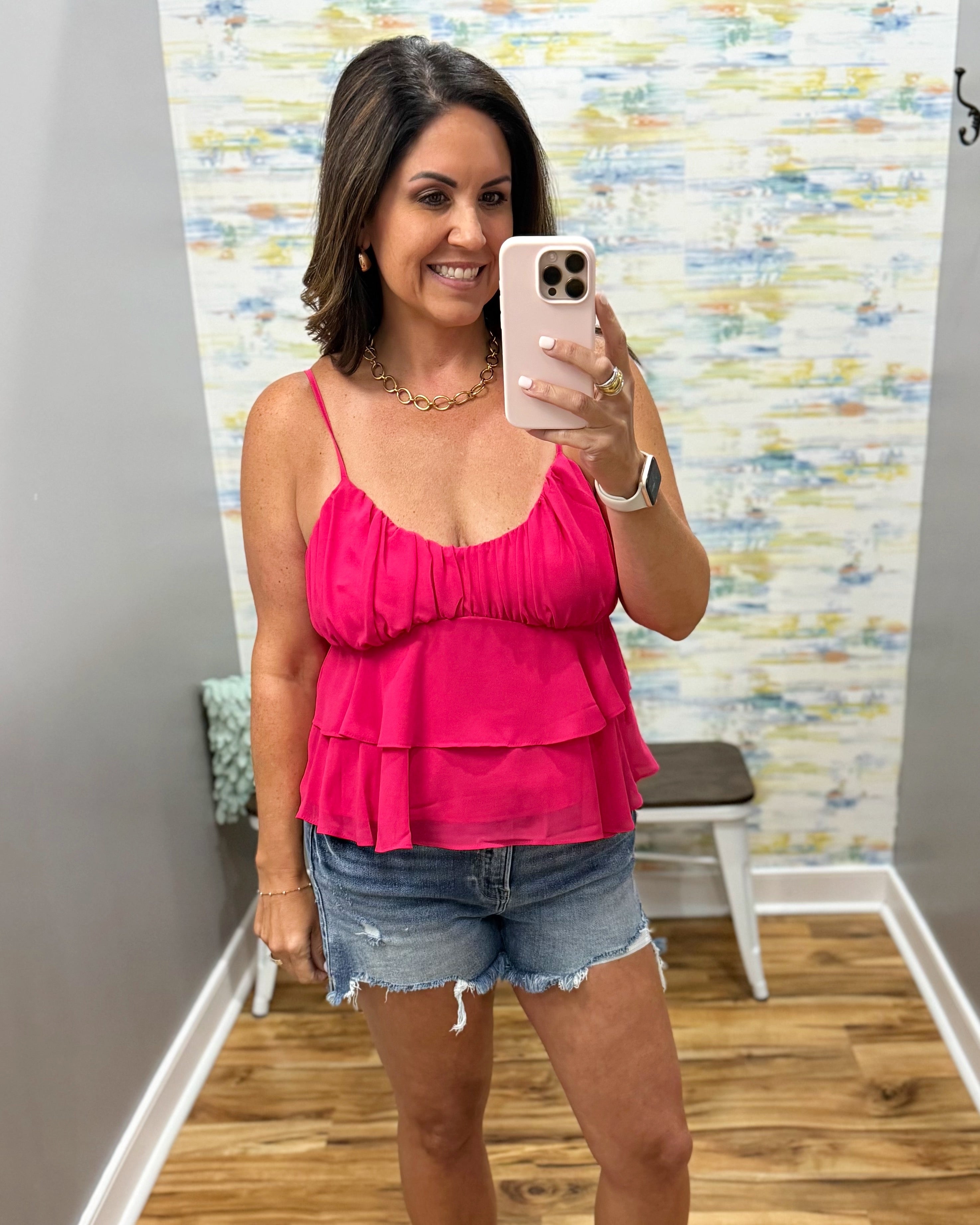 Woman taking a mirror selfie wearing a pink top and denim shorts in a room with a colorful wall.
