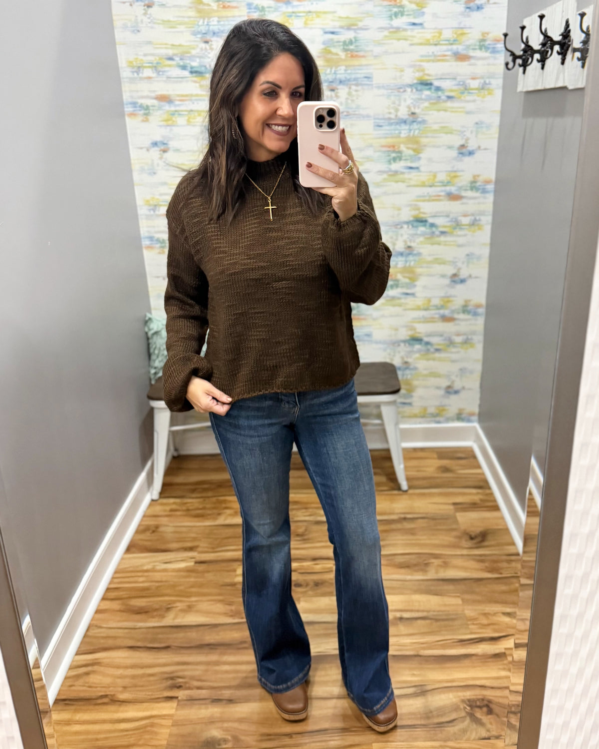 A woman takes a mirror selfie in a fitting room wearing a brown textured knit sweater, blue flare jeans, and brown boots.
