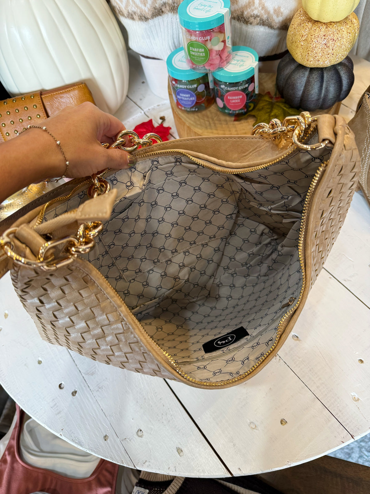 A woven beige shoulder bag with gold-tone hardware and a patterned interior lining, held open by a hand over a white wooden table.