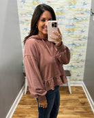 A woman takes a mirror selfie wearing a dusty rose long-sleeve cropped hoodie with a ruffled hem and dark blue jeans.
