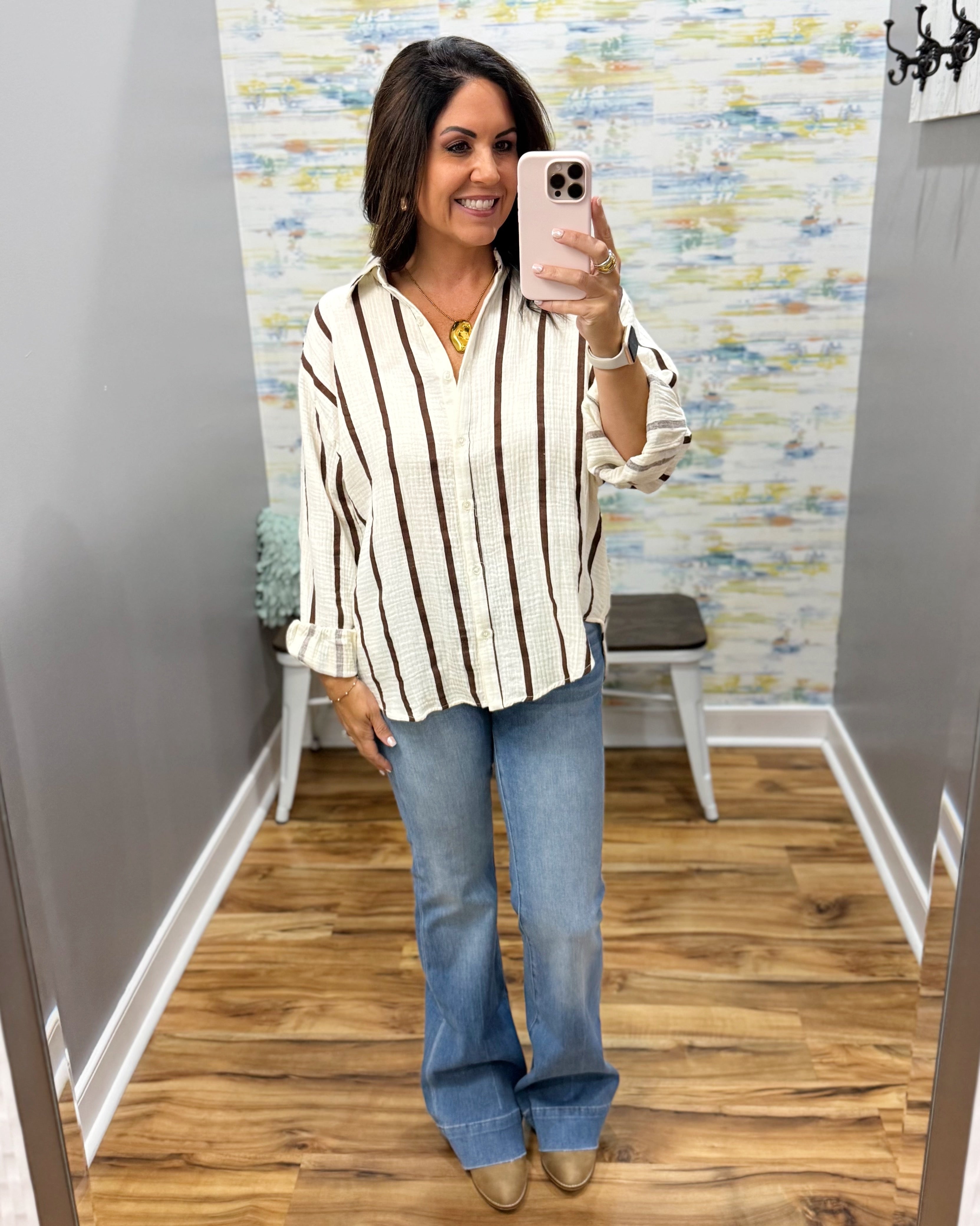 A woman takes a mirror selfie in a fitting room wearing a cream and brown vertically striped button-down shirt, blue flare jeans, and tan shoes.