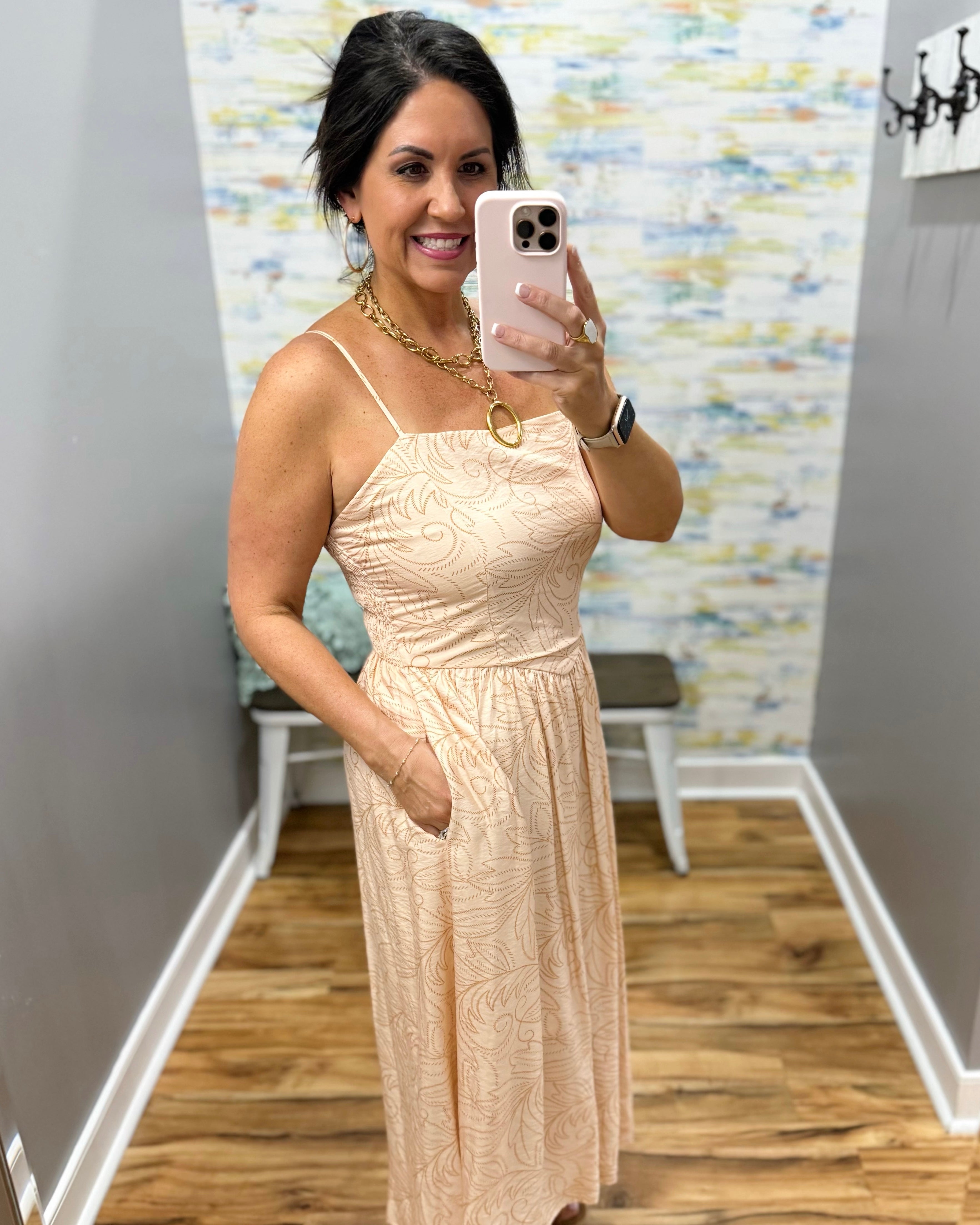 Woman taking a mirror selfie wearing a beige dress in a room with patterned wallpaper.
