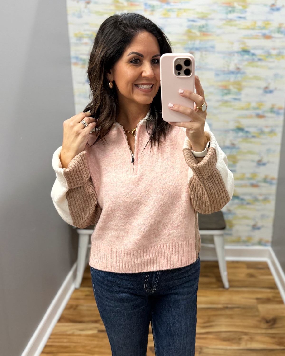 A woman takes a mirror selfie in a fitting room wearing a light pink quarter-zip sweater with contrasting cream and tan knit sleeves, paired with dark blue jeans.