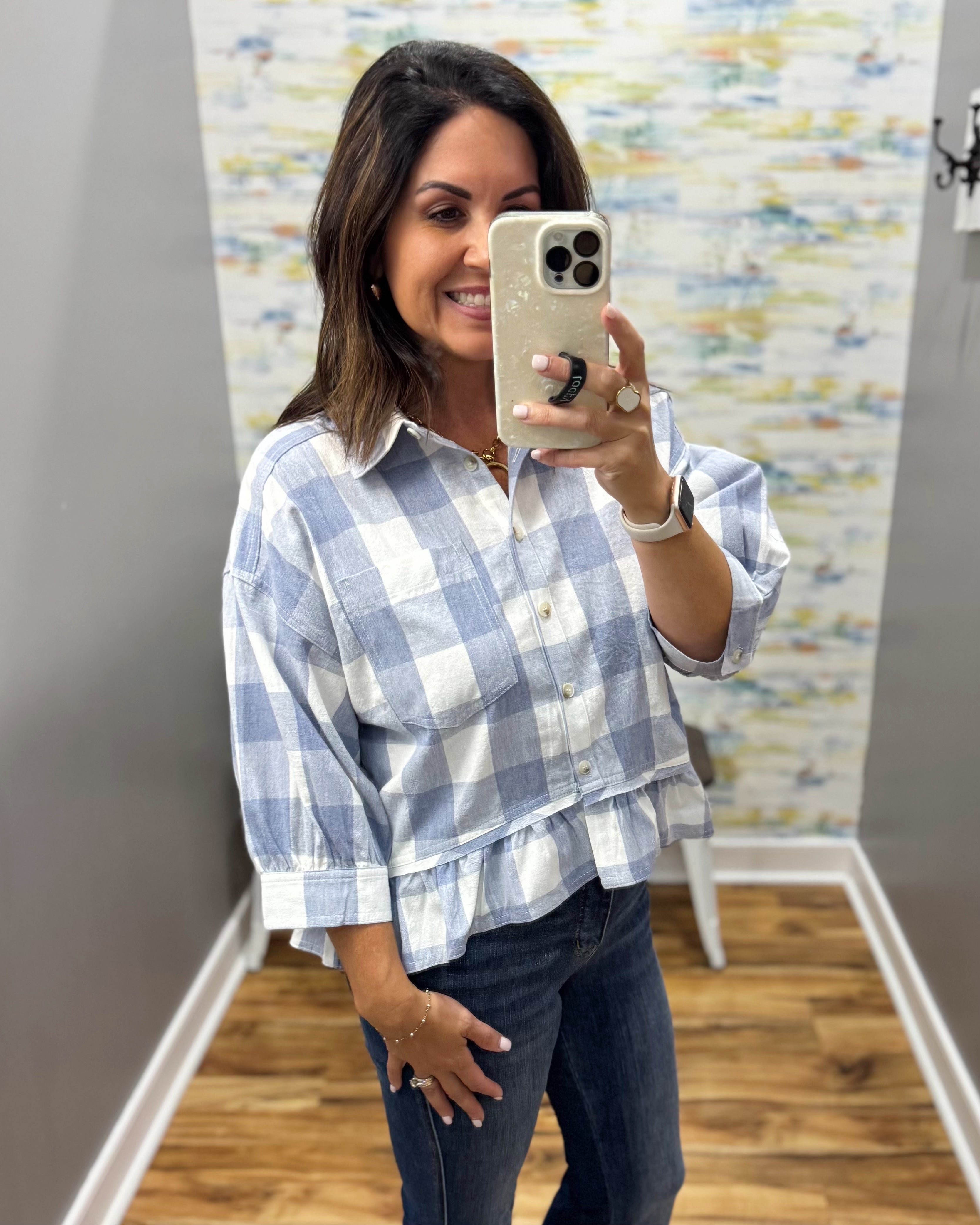 A woman takes a mirror selfie wearing a light blue and white gingham check long-sleeve button-up shirt with dark blue jeans.