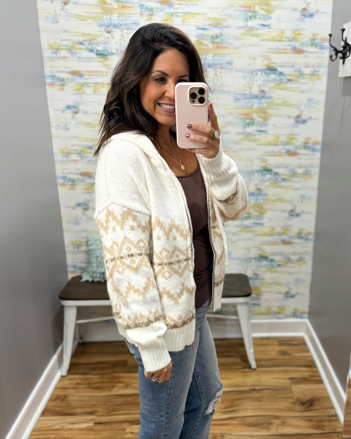 A woman takes a mirror selfie wearing a cream and beige patterned knit cardigan over a brown top and blue jeans, standing in a room with patterned wallpaper and a wooden floor.