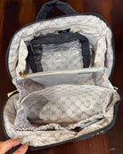 An open makeup bag or organizer shown from above on a dark wooden surface. The exterior is dark gray/black, while the interior is lined with a beige and gray repeating monogram pattern.