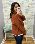 A woman takes a mirror selfie in a boutique showing a rust-colored knit sweater with a white scalloped hem and cuffs, paired with blue jeans.