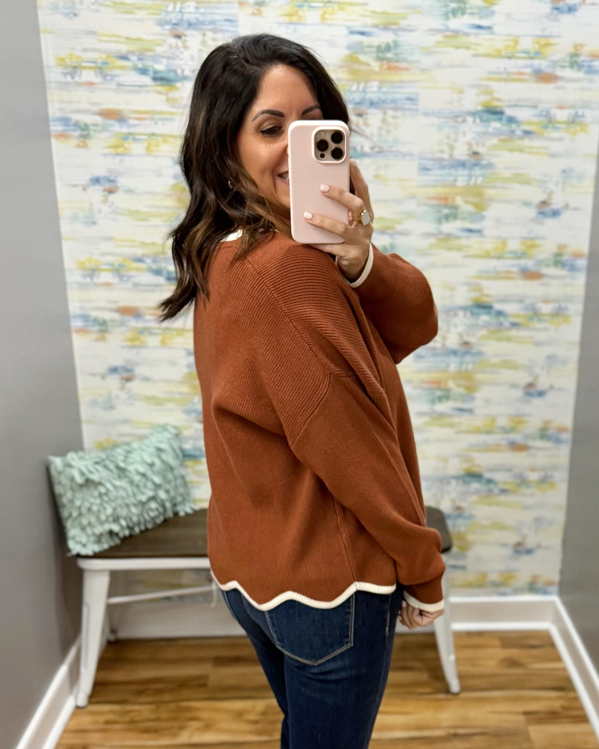 A woman takes a mirror selfie in a boutique showing a rust-colored knit sweater with a white scalloped hem and cuffs, paired with blue jeans.