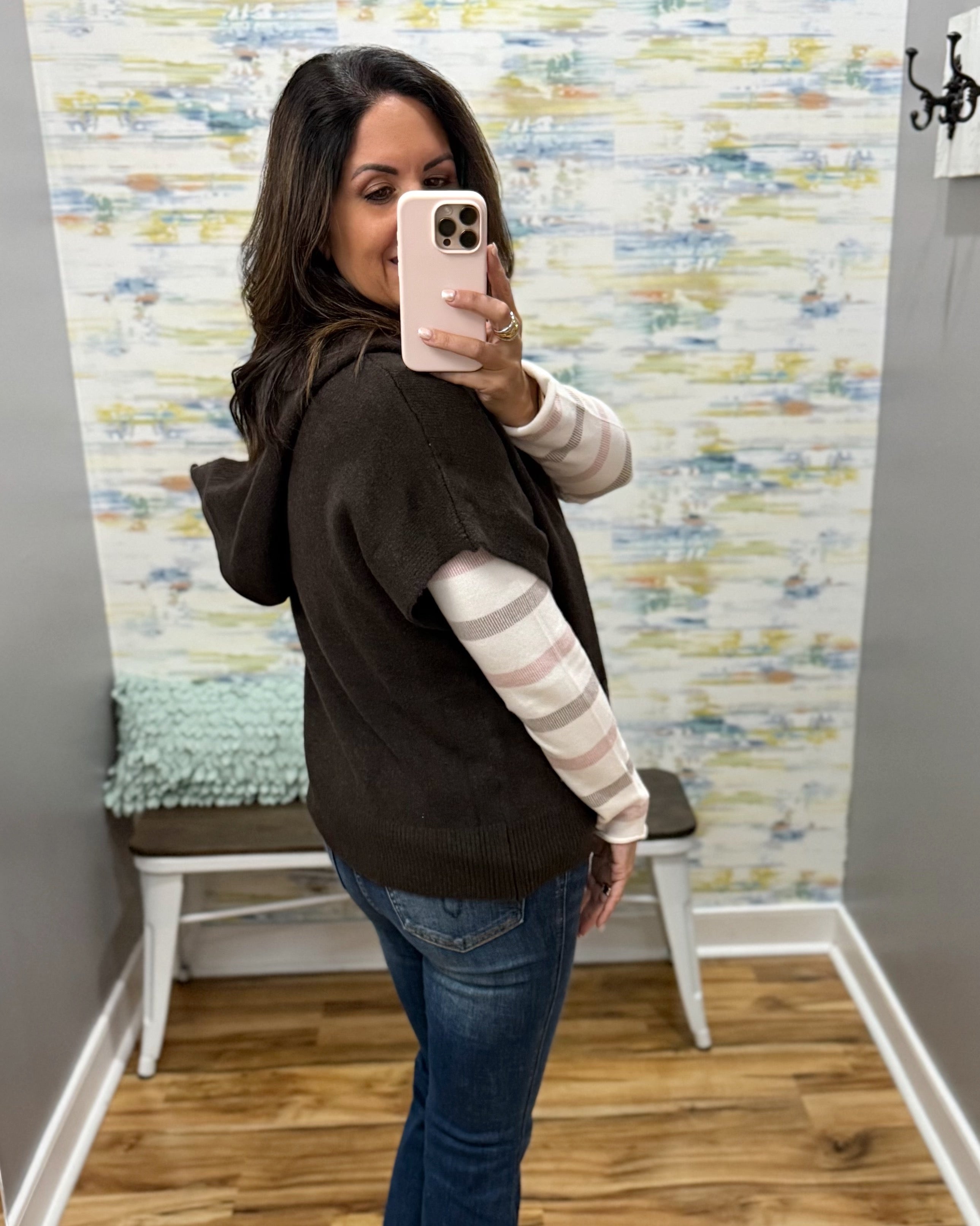 A woman takes a mirror selfie wearing a dark brown knit hoodie vest over a pink and white striped long-sleeve shirt, paired with blue jeans, in a room with patterned wallpaper and a wooden floor.