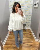 A woman takes a mirror selfie wearing a cream-colored textured hoodie, blue jeans, and tan boots in a home hallway.