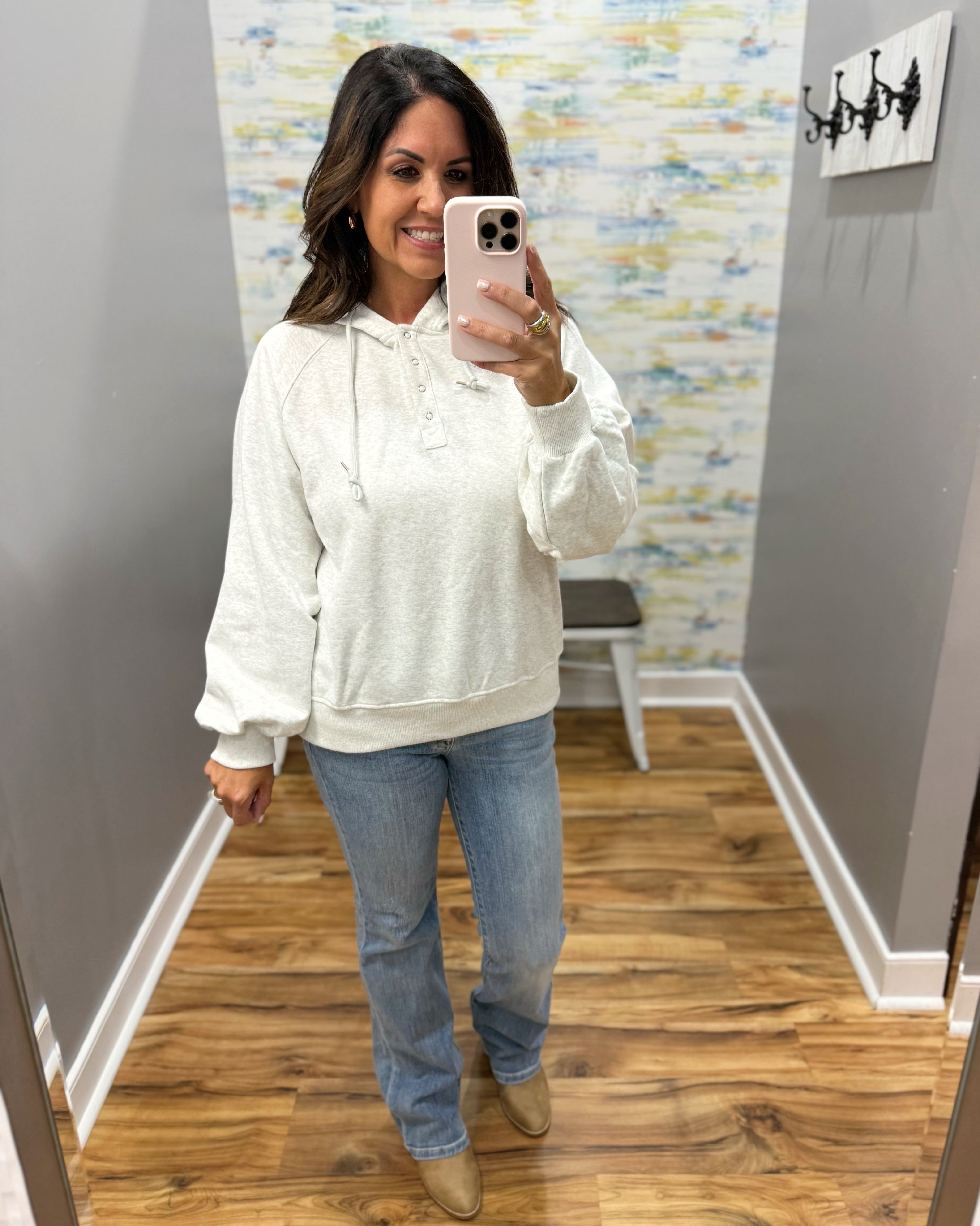 A woman takes a mirror selfie wearing a cream-colored textured hoodie, blue jeans, and tan boots in a home hallway.
