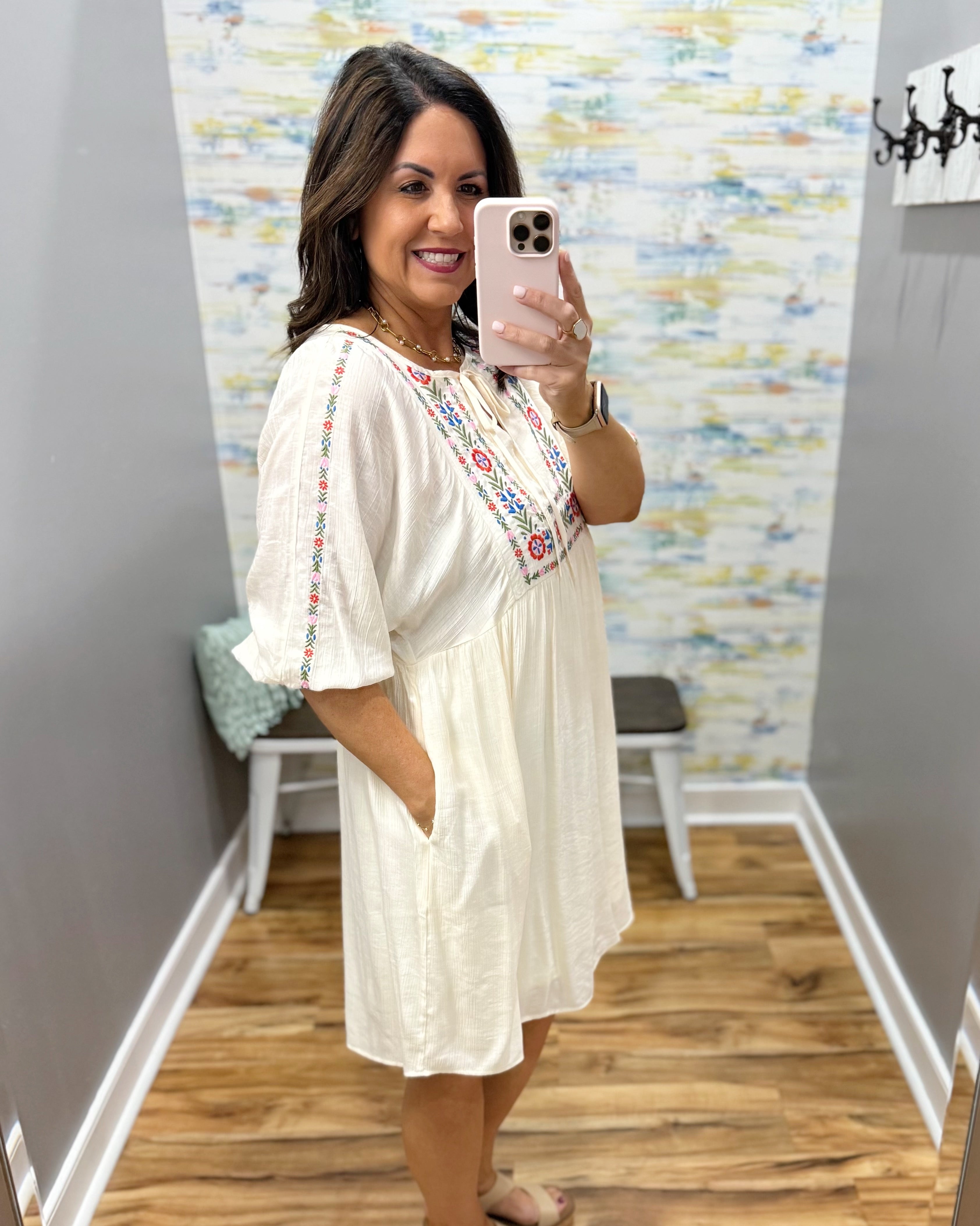Woman taking a mirror selfie wearing a white dress with colorful embroidery in a room.