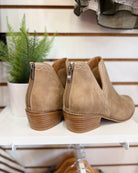 A pair of beige suede ankle boots with side zippers and stacked block heels, displayed on a white shelf next to a potted green plant.