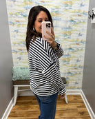 A woman takes a mirror selfie wearing a black and white horizontally striped long-sleeve top and blue jeans in a fitting room with a patterned wallpaper background.