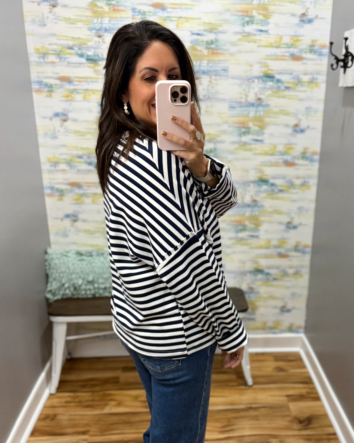 A woman takes a mirror selfie wearing a black and white horizontally striped long-sleeve top and blue jeans in a fitting room with a patterned wallpaper background.
