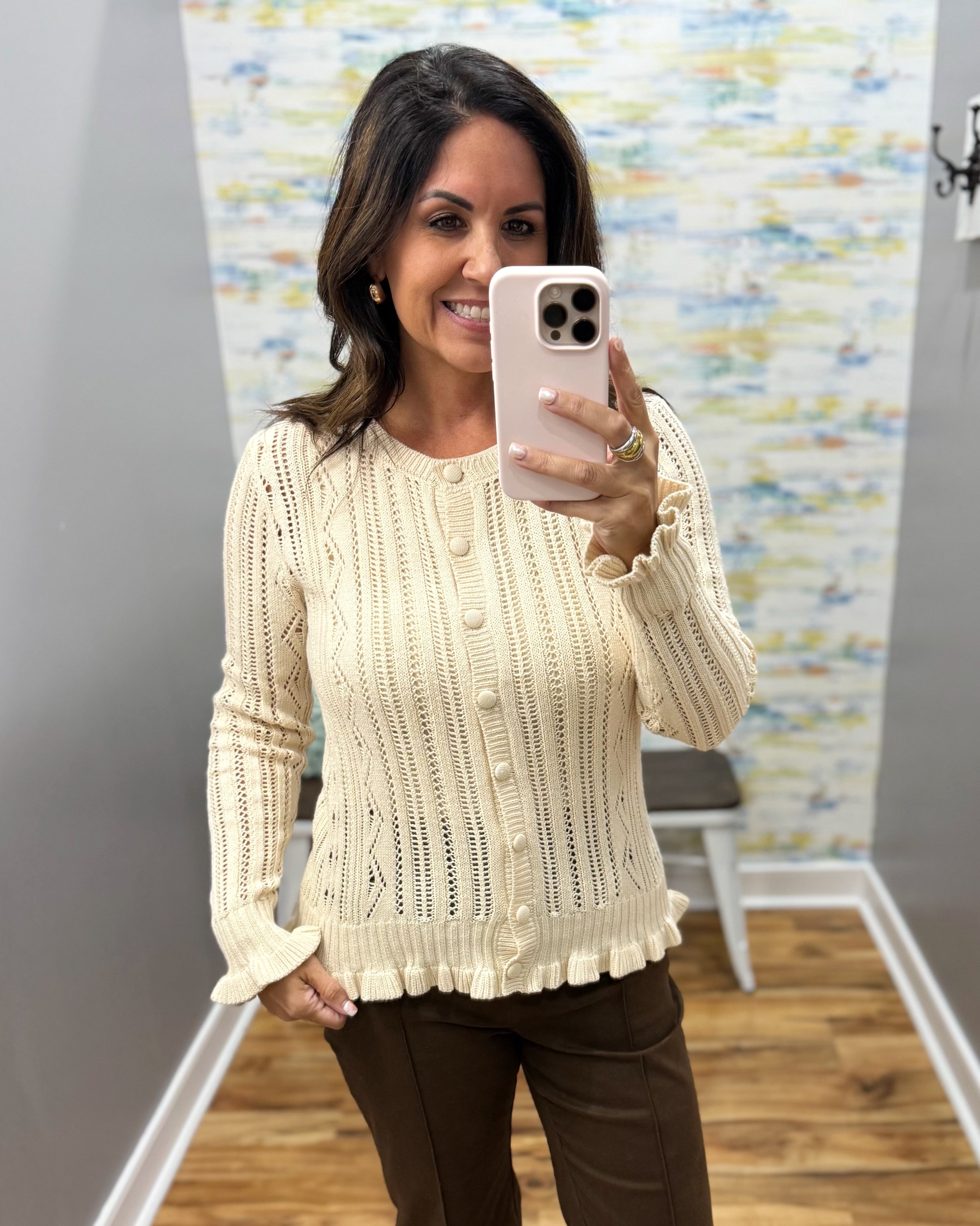 A woman takes a mirror selfie wearing a cream cable-knit button-front sweater with ruffled cuffs and hem, paired with dark brown pants, in front of a patterned wall.