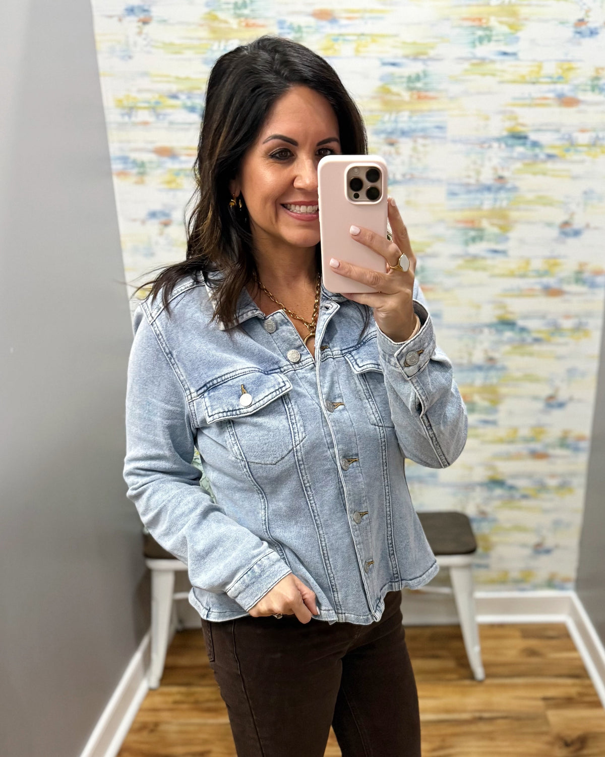 A woman takes a mirror selfie wearing a light-wash blue denim jacket and dark brown pants in an indoor room with a patterned wall and wood floor.