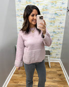 A woman takes a mirror selfie wearing a light pink knit sweater and gray pants in a room with a wood floor and a colorful abstract-patterned wall.
