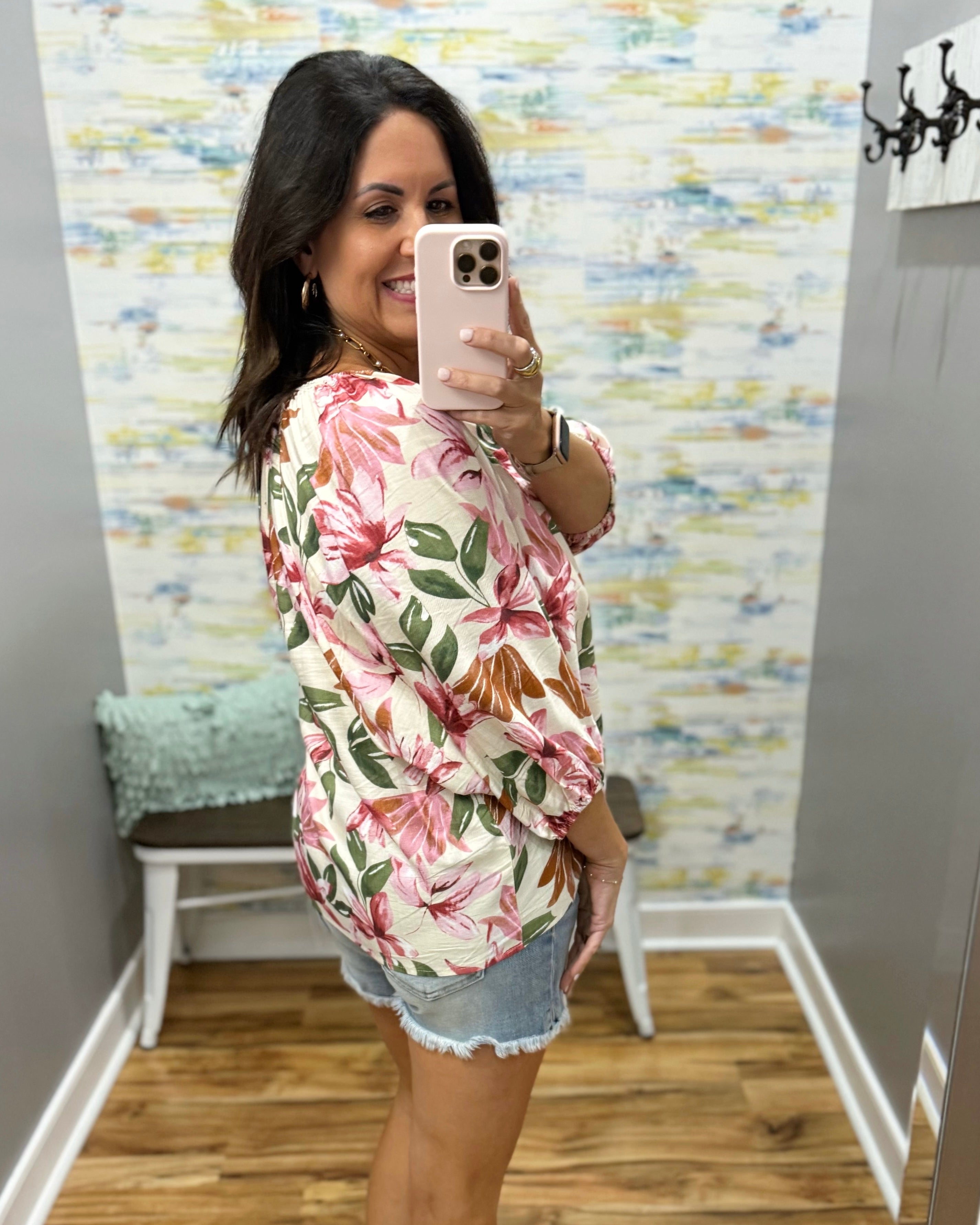 Woman taking a mirror selfie wearing a floral shirt and denim shorts in a room with colorful wall art.