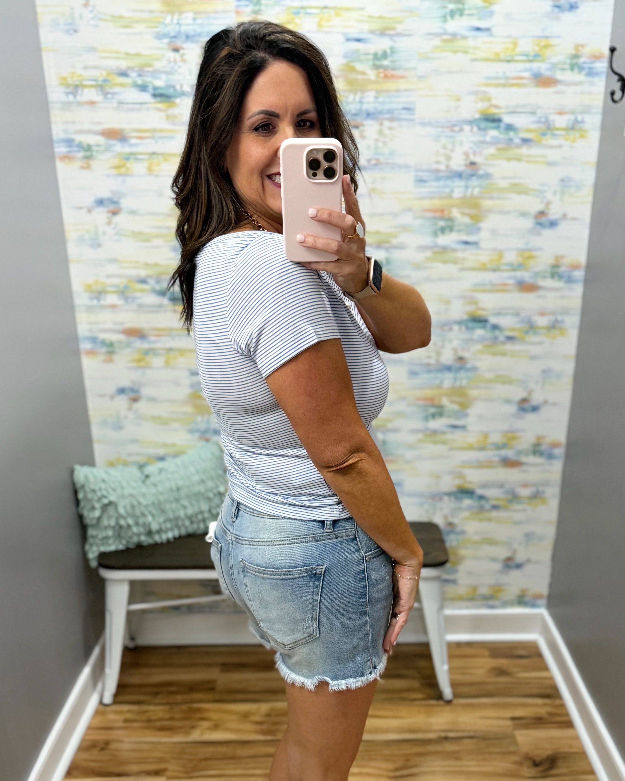 Woman taking a mirror selfie wearing a striped shirt and denim shorts in a room with a colorful wall.