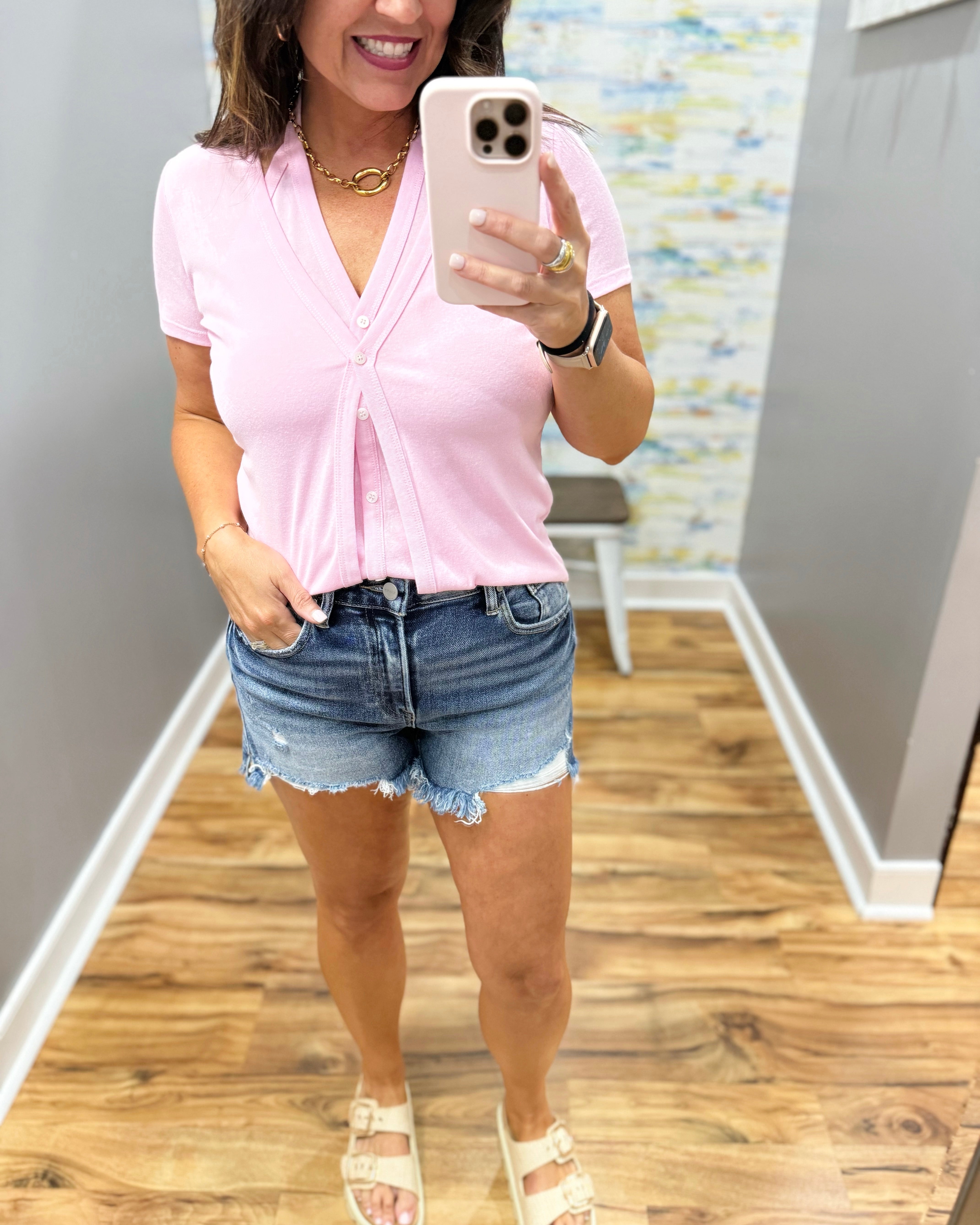 Woman taking a mirror selfie wearing a pink top and denim shorts indoors.