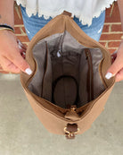 A tan bucket bag with a gold-tone clasp, held open by a person's hands to show the interior lining and zip compartments.