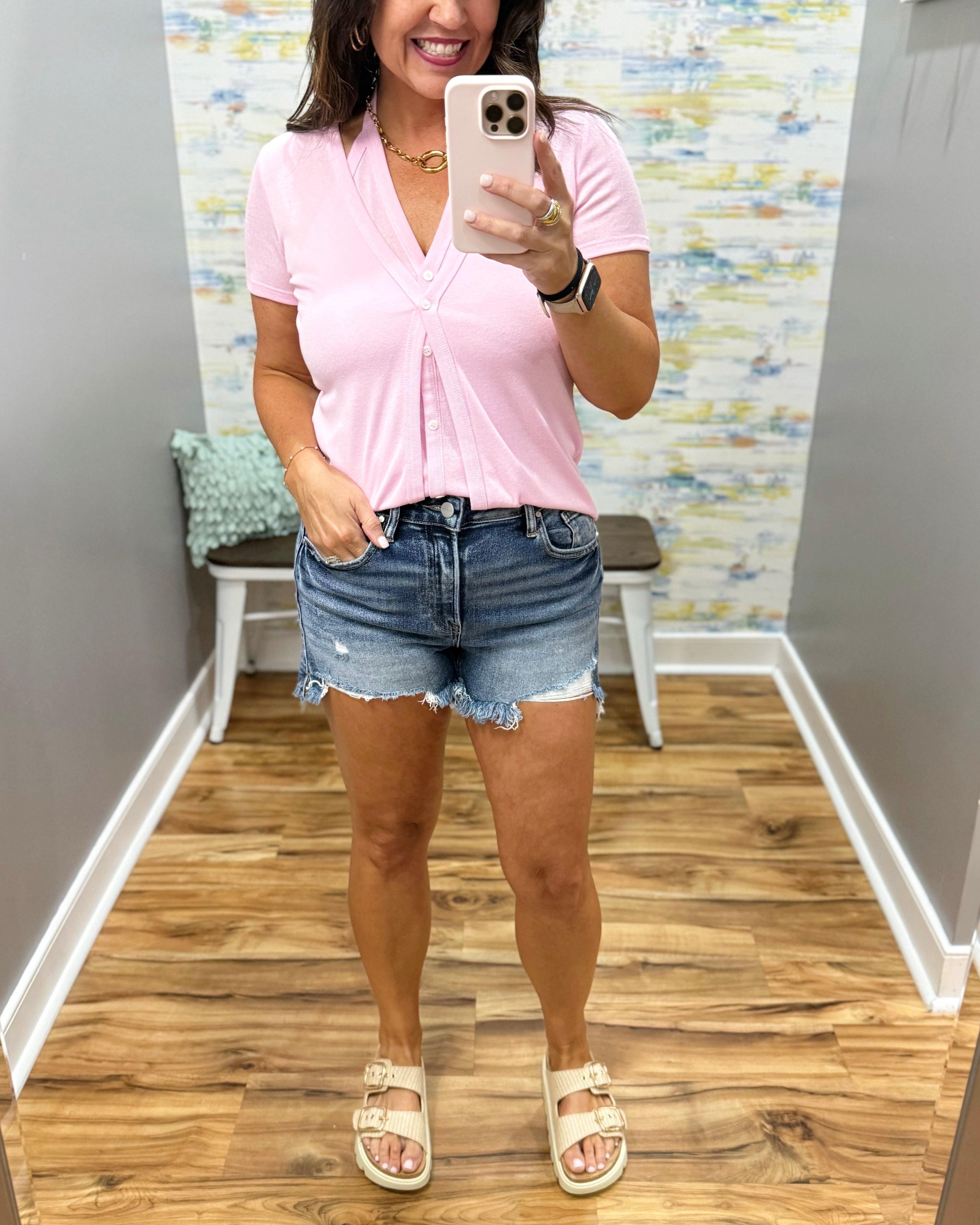 Woman taking a mirror selfie wearing a pink shirt and denim shorts in a room with wooden flooring.