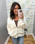 A woman taking a mirror selfie wearing a cream and tan Fair Isle pattern zip-up cardigan over a brown top and blue jeans.