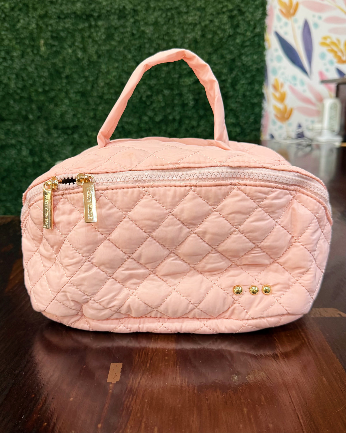 A light pink quilted cosmetic bag with a top handle, gold-tone zipper pulls, and small gold studs on the front, displayed on a dark wooden surface.