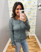 A woman takes a mirror selfie wearing a slate blue-gray cable-knit button-up cardigan sweater and light-wash blue jeans in a home setting with wood floors and a patterned wall.