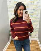 A woman takes a mirror selfie wearing a ribbed mock neck sweater with horizontal stripes in burgundy and orange, paired with blue jeans.