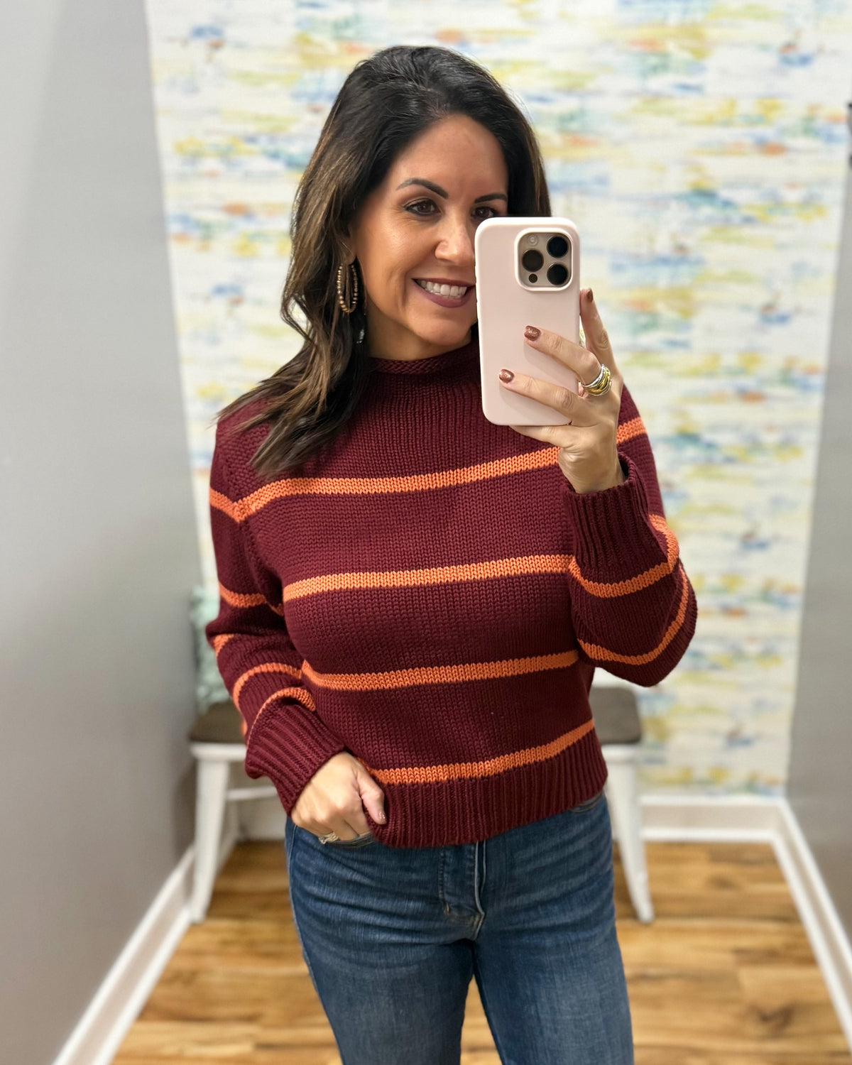 A woman takes a mirror selfie wearing a ribbed mock neck sweater with horizontal stripes in burgundy and orange, paired with blue jeans.