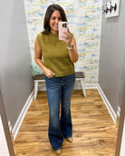 A woman takes a mirror selfie in a fitting room wearing a sleeveless olive green knit top and dark blue flare jeans.