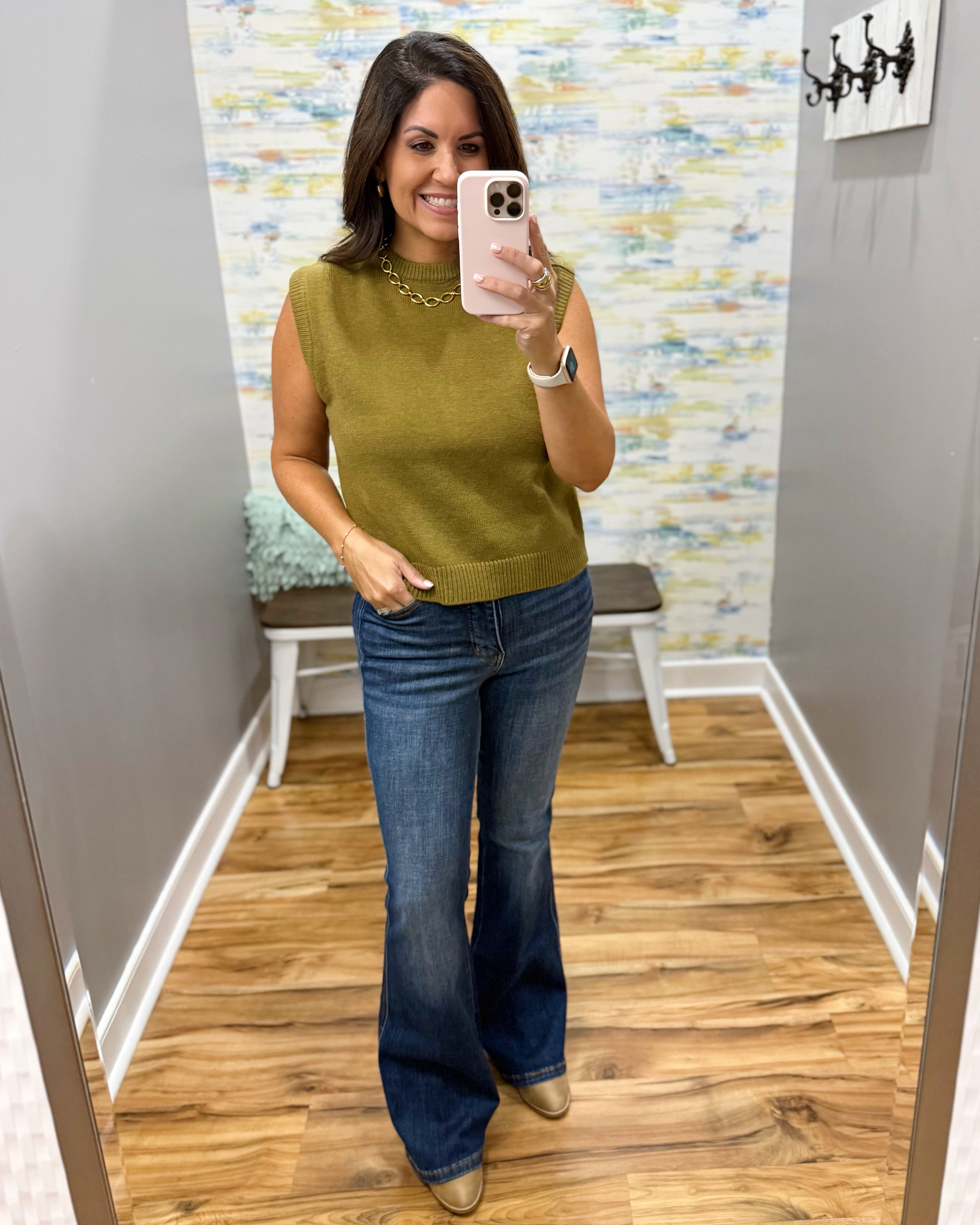 A woman takes a mirror selfie in a fitting room wearing a sleeveless olive green knit top and dark blue flare jeans.