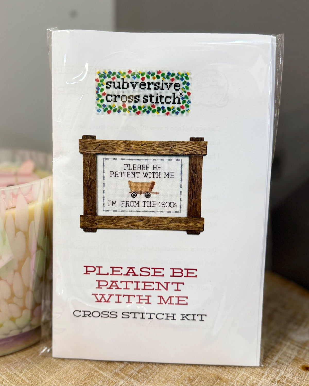 A packaged cross stitch kit labeled 'subversive cross stitch' with the design text 'PLEASE BE PATIENT WITH ME I'M FROM THE 1900s' shown on the front, photographed standing upright on a fabric surface.