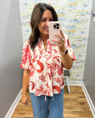 Woman taking a mirror selfie wearing a red floral blouse and blue jeans in a room with a colorful wall.
