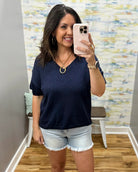 Woman taking a mirror selfie wearing a navy blue t-shirt and light denim shorts in a room with patterned wallpaper.