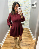 A woman takes a mirror selfie wearing a long-sleeve burgundy mini dress with a zip-up front and beige knee-high boots, standing in a hallway with wood floors.