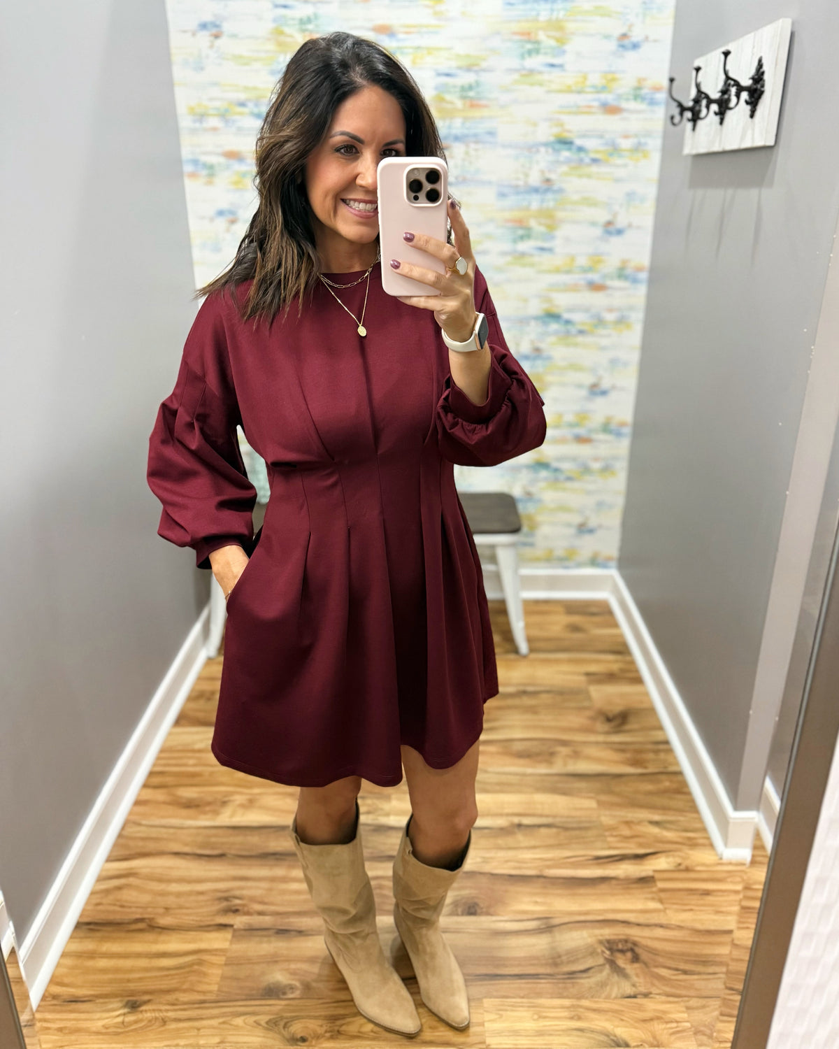 A woman takes a mirror selfie wearing a long-sleeve burgundy mini dress with a zip-up front and beige knee-high boots, standing in a hallway with wood floors.