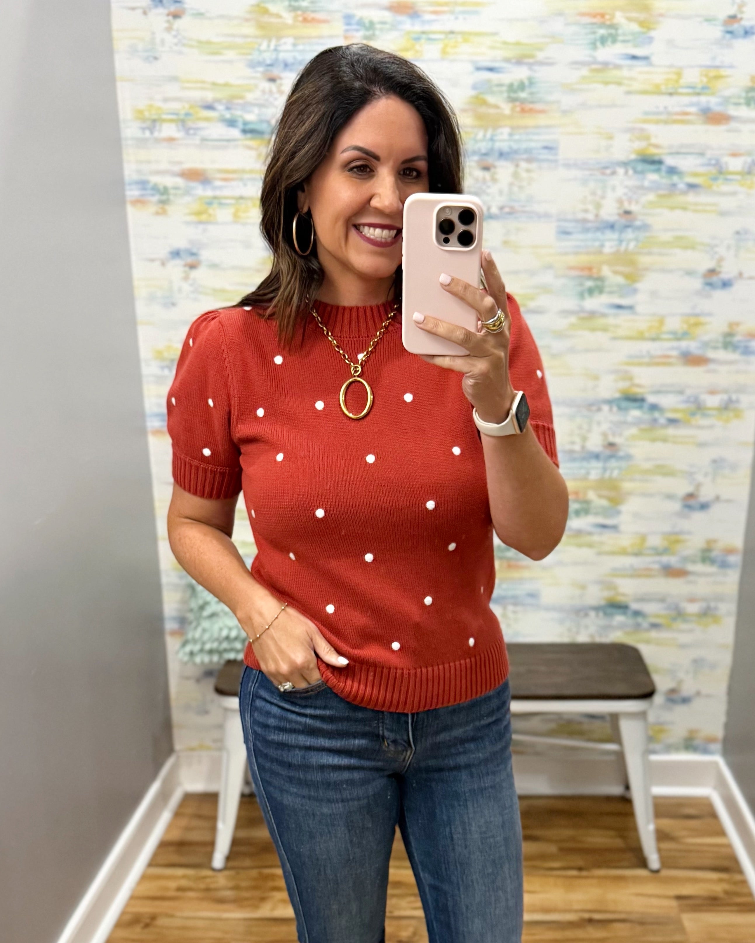 A woman takes a mirror selfie wearing a rust-red short-sleeve knit sweater with white polka dots and blue jeans, standing in front of a patterned wallpapered wall.
