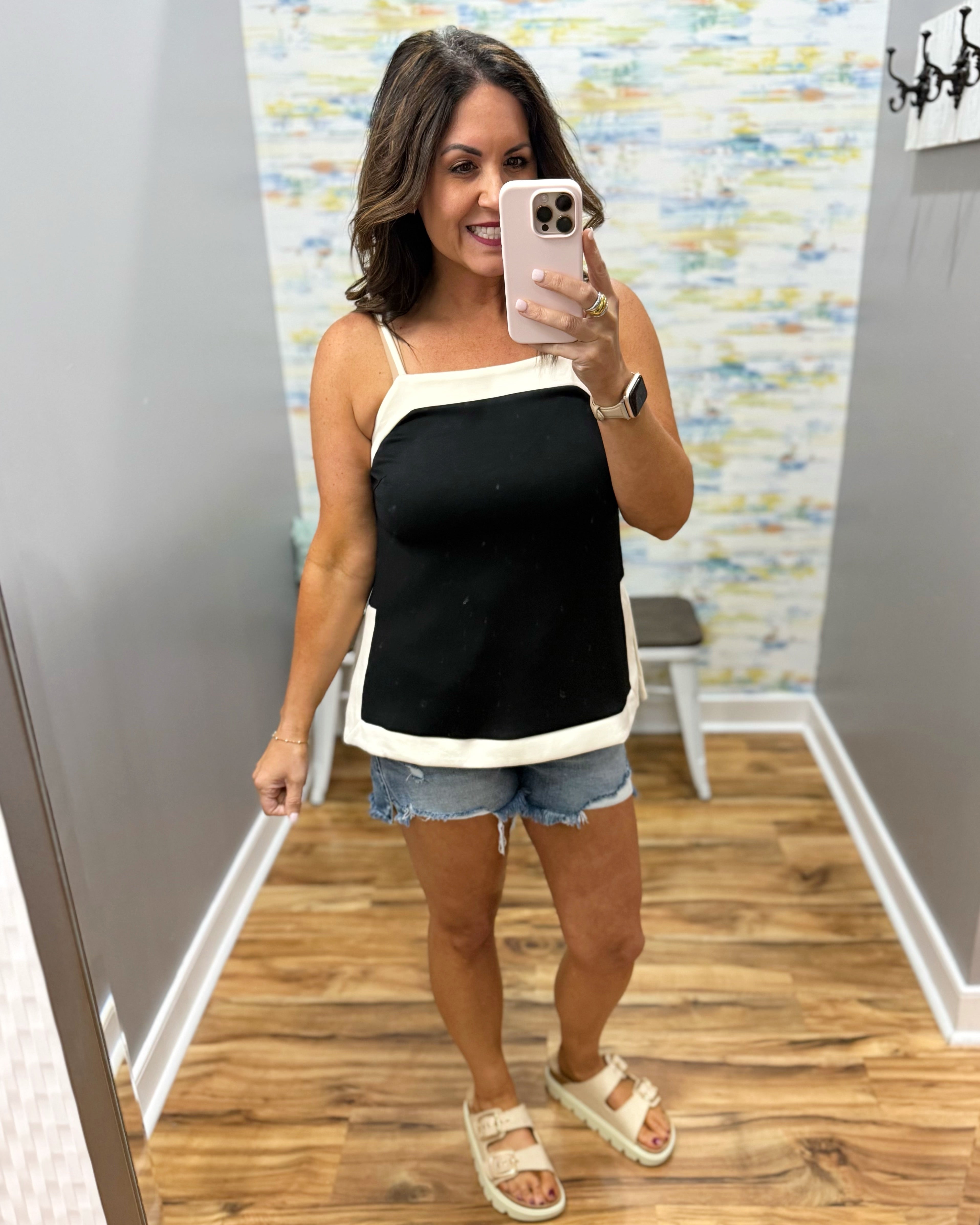 Woman taking a mirror selfie wearing a black and white top and denim shorts in a room with patterned wallpaper.