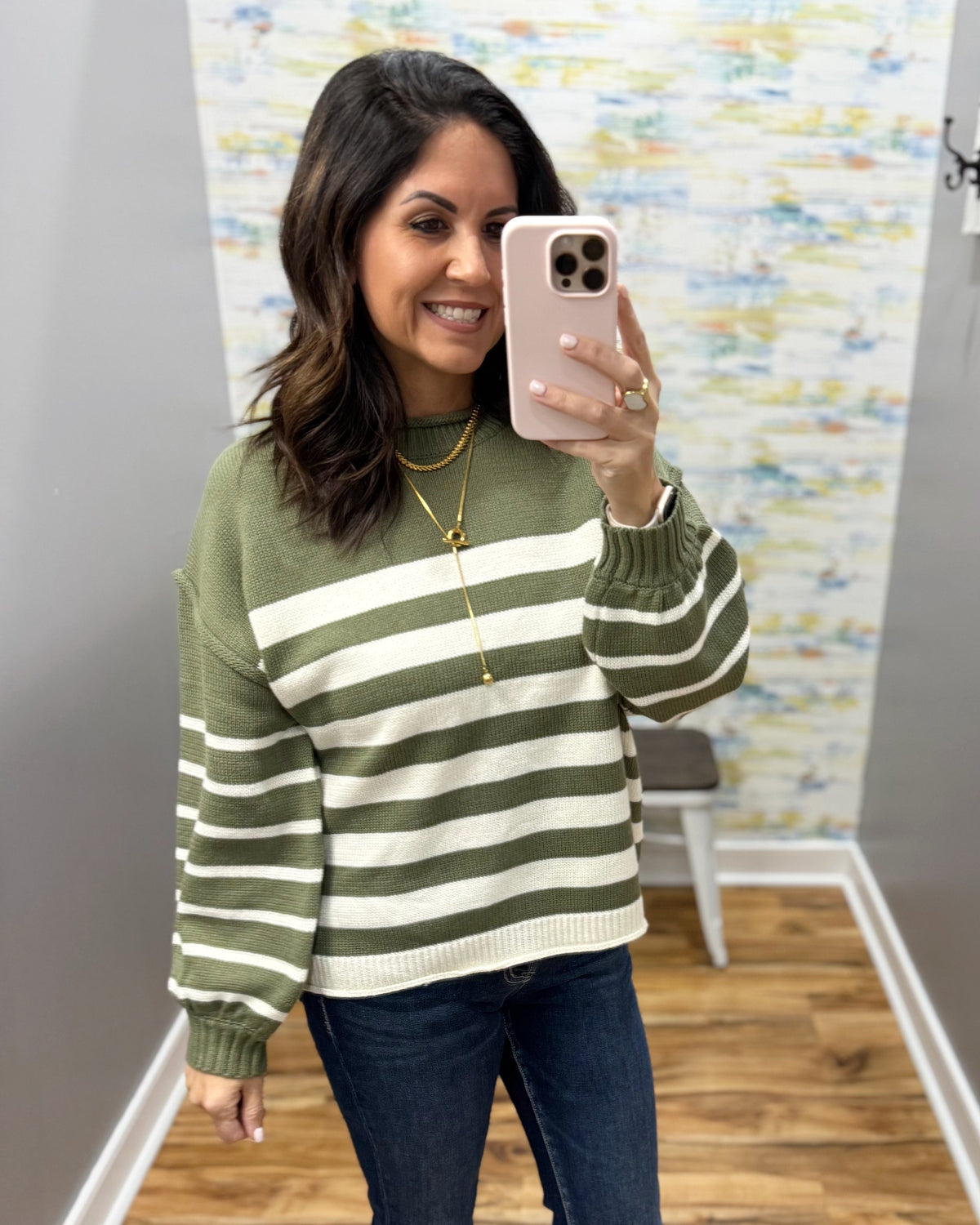 A woman takes a mirror selfie wearing an olive green and white horizontally striped knit sweater, dark blue jeans, and a gold necklace in what appears to be a boutique fitting room.