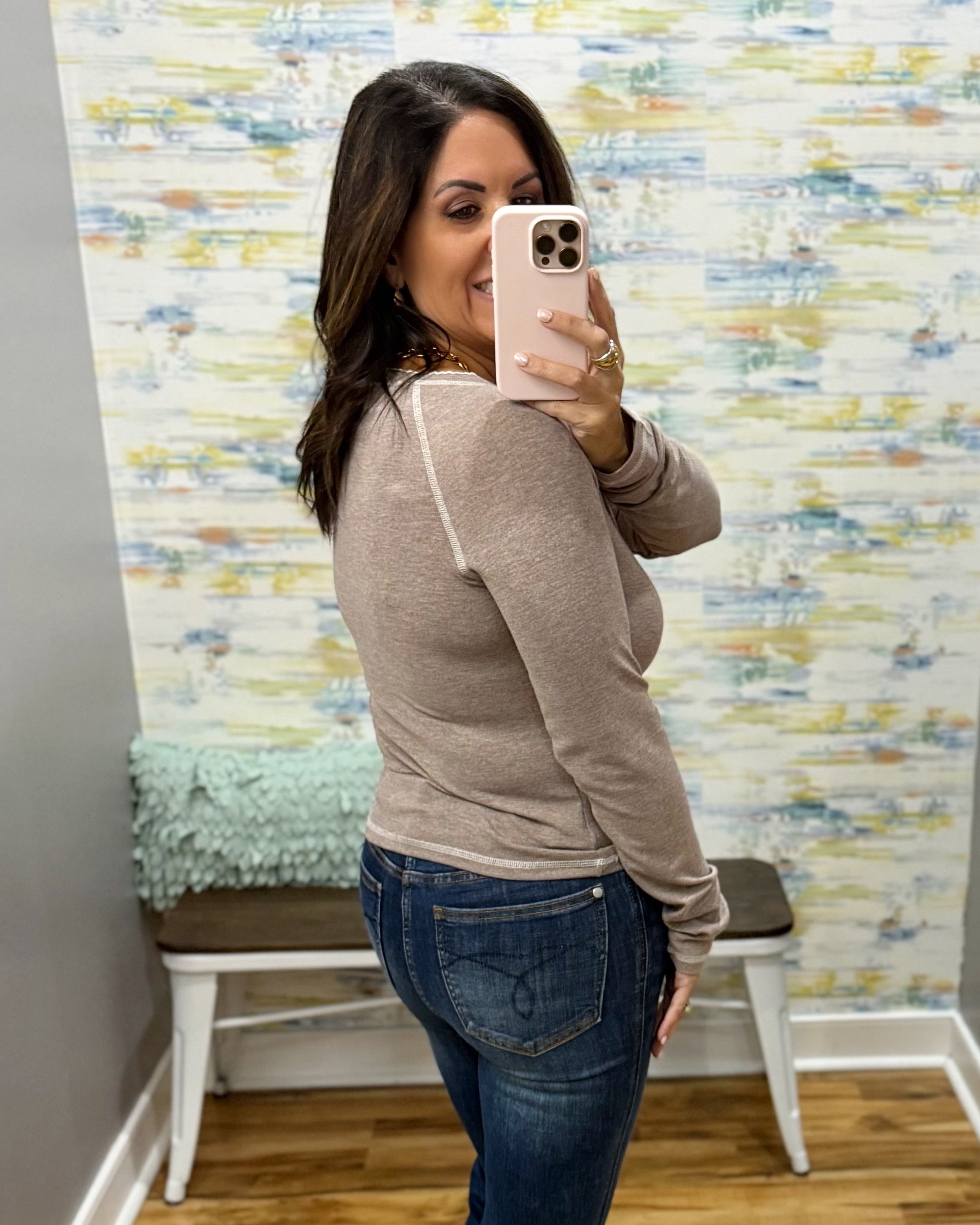 A woman takes a mirror selfie in a fitting room wearing a taupe long-sleeve knit top and blue jeans. The background features a colorful abstract patterned wallpaper and a small white bench.