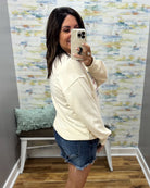 A woman takes a mirror selfie wearing a cream-colored oversized sweatshirt and denim shorts in a fitting room with a patterned wall and wooden floor.