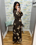 A woman takes a mirror selfie in a boutique fitting room wearing a long-sleeve brown maxi dress with a beige floral pattern. The room features a colorful abstract wallpaper, a wooden floor, and a small bench with a mint green pillow.