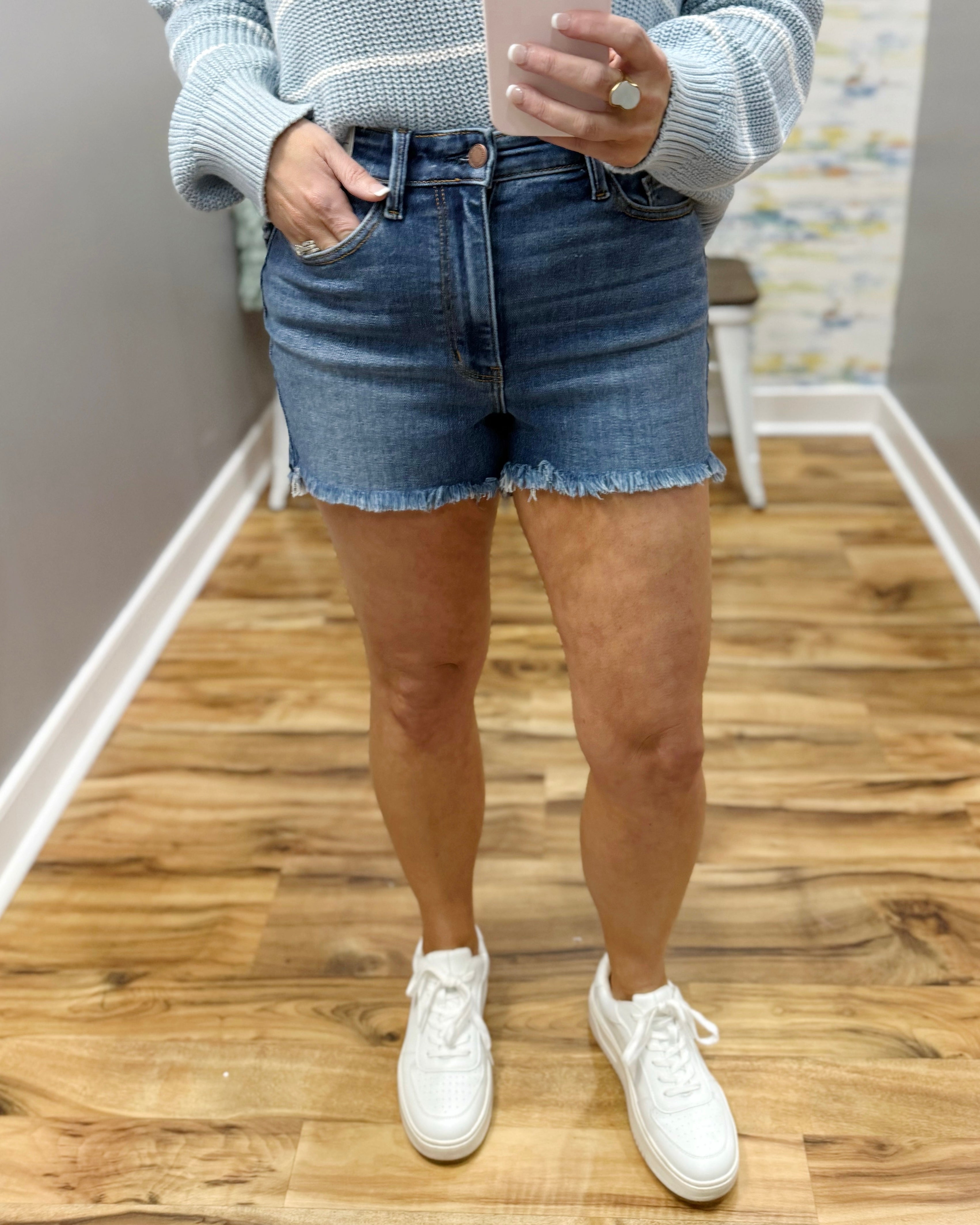 Person wearing a light gray sweater, blue denim shorts, and white sneakers on a wooden floor.