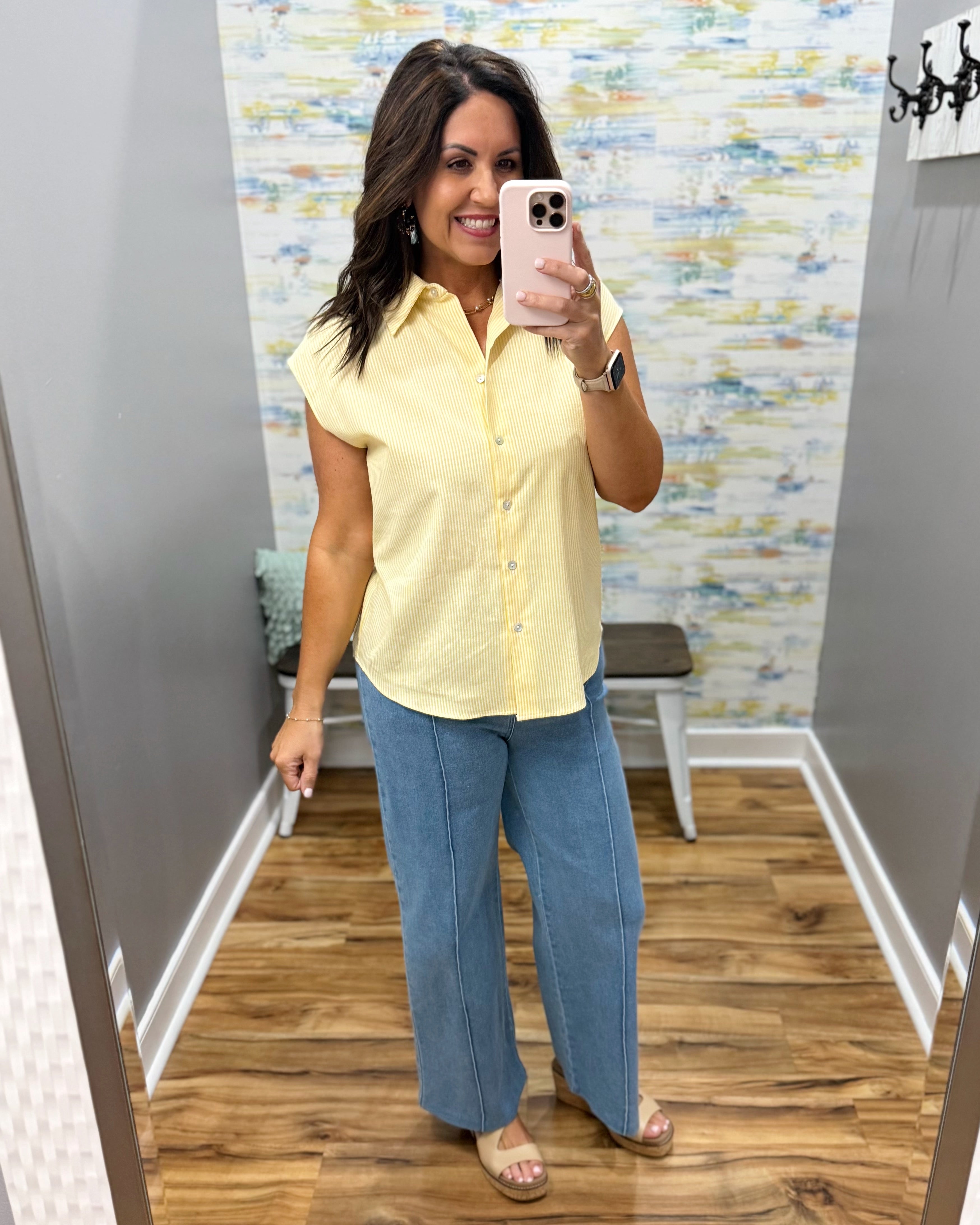 Woman taking a mirror selfie wearing a yellow shirt and blue jeans in a room with patterned wallpaper.