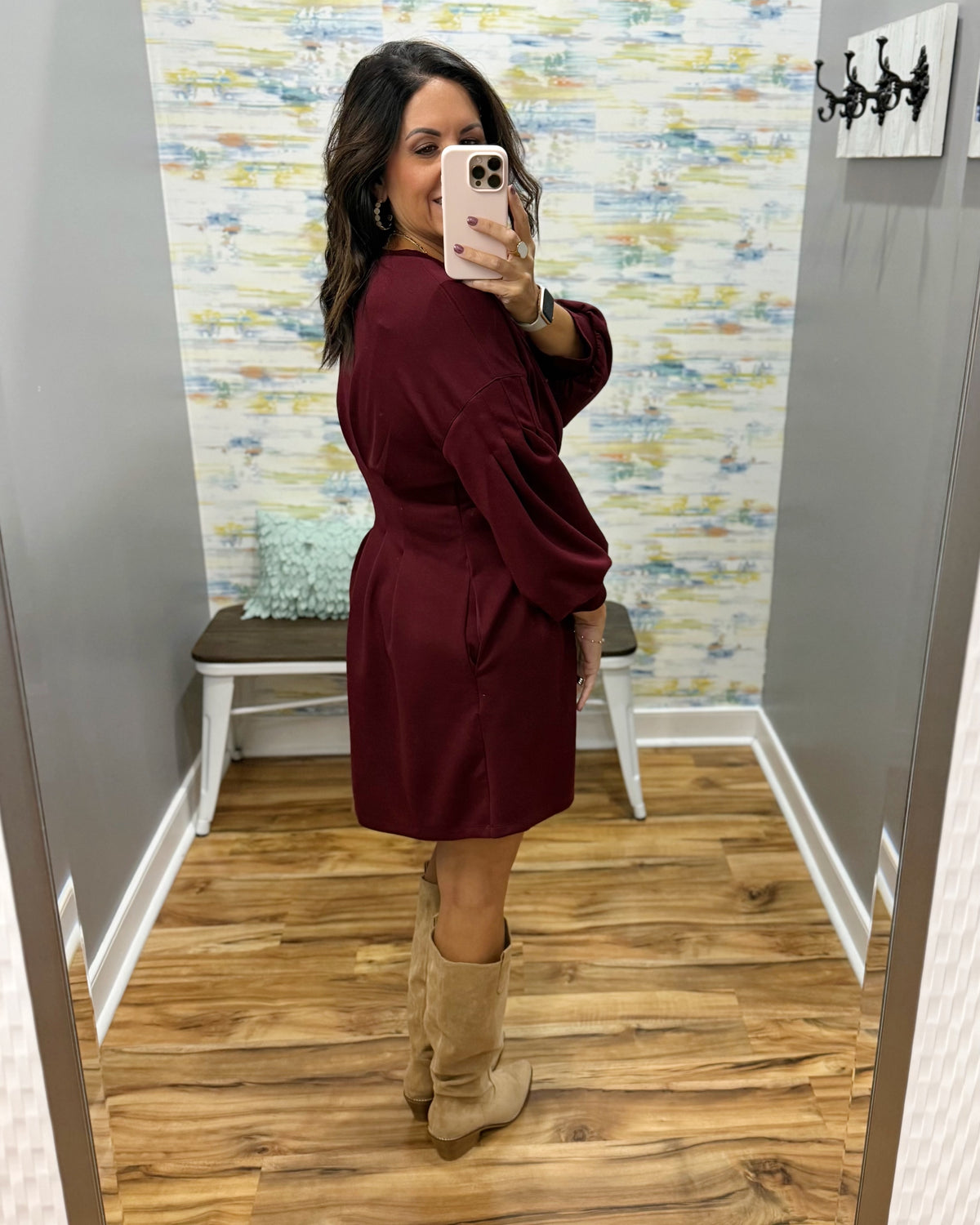 A woman takes a mirror selfie in a fitting room wearing a long-sleeve burgundy wrap-style dress with beige suede boots. The background features gray walls, a patterned wallpaper, a wooden floor, and a small bench.