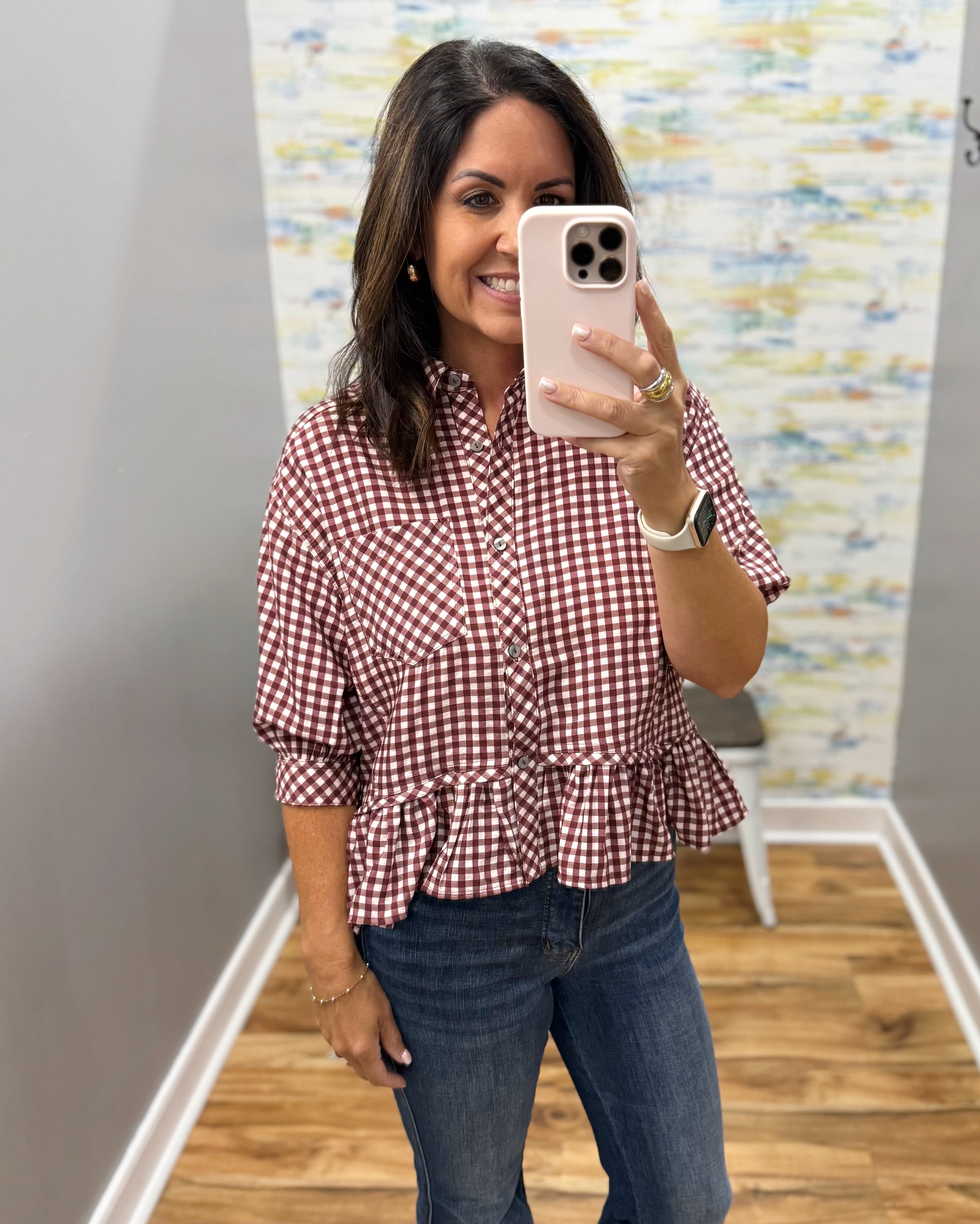 A woman taking a mirror selfie wearing a red and white gingham check long-sleeve blouse with a ruffled hem and blue jeans.